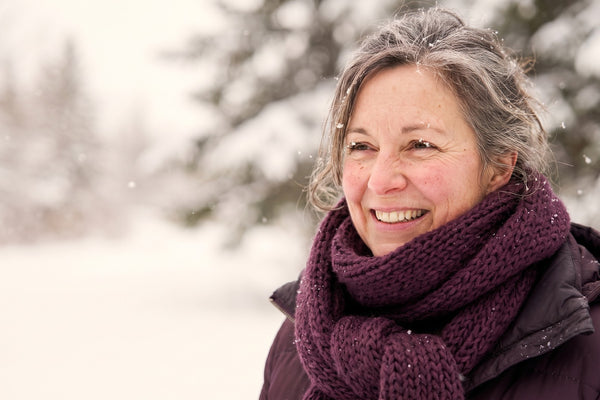 Femme québécoise d’une cinquantaine d’années souriante dehors sous la neige, écharpe aubergine, flocons sur les cils