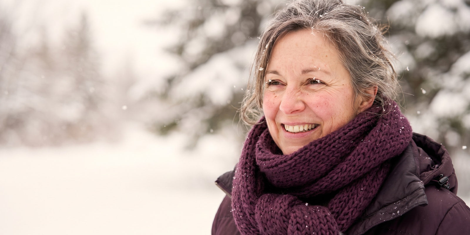 Femme québécoise d’une cinquantaine d’années souriante dehors sous la neige, écharpe aubergine, flocons sur les cils
